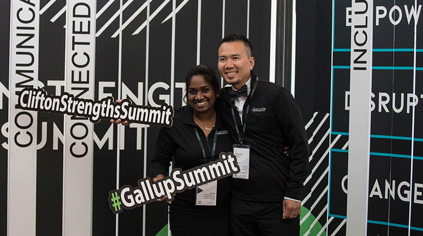 Two employees taking a picture hold a CliftonStrengthsSummit sign and a #GallupSummit sign.