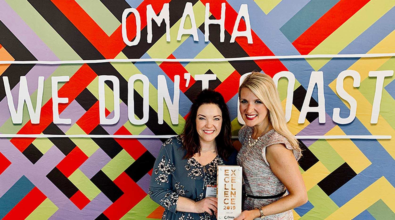 Two employees showing off an Excellence 2019 program in front of a sign saying 'Omaha We don't Coast'