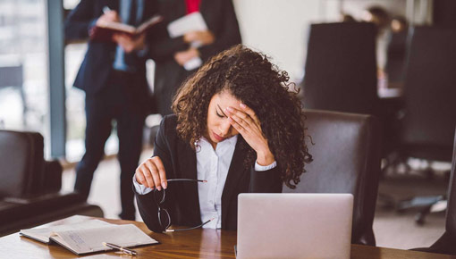 A young woman in a suit jacket rests her head on her forehead in frustration while seated at her office desk