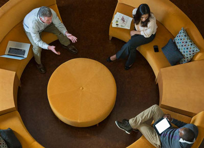 An overhead view of several coworkers sitting on a circular couch with laptop computers, talking.