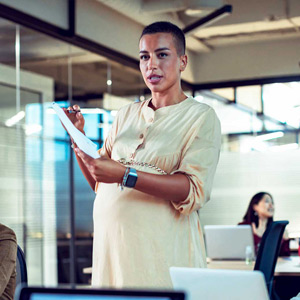 A pregnant woman stands to present with papers in her hand in a brightly lit office.