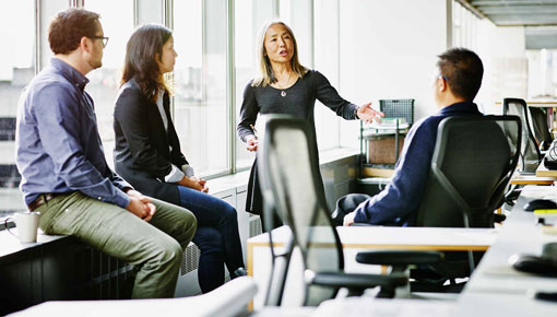 A woman gestures with her arm and speaks to a group of coworkers gathered around her in a brightly lit office.