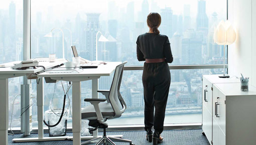 A woman looks out an office window at a city, seen from behind