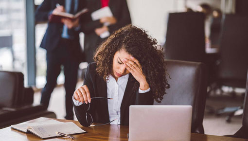 A woman rests her forehead against her hand while seated at a work desk in front of a laptop computer