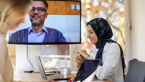 A smiling woman wearing a headscarf works on a laptop computer in front of a man on a video screen