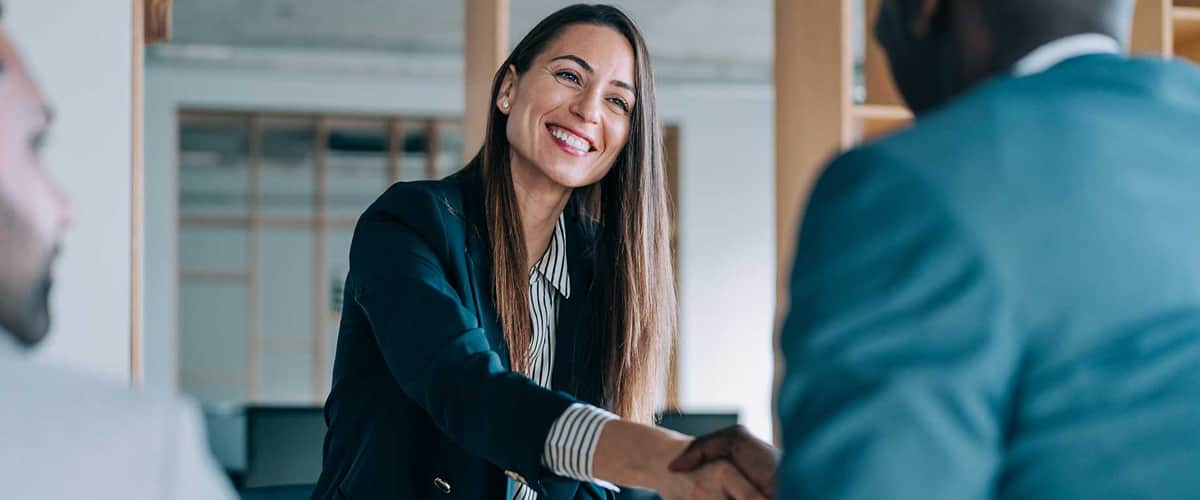 A smiling woman in a suit shakes hands with a man in an office.