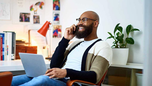 Smiling man talks on the phone with a laptop on his lap in a home office