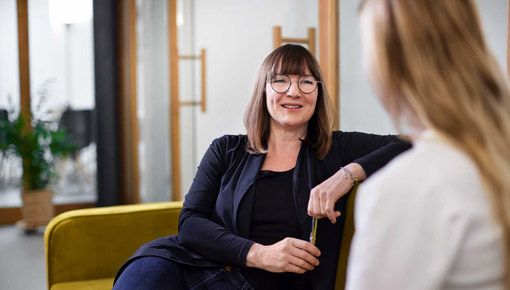 A smiling woman in glasses sits in an armchair and talks to another woman, whose back is to the viewer.