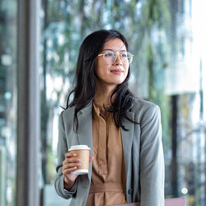 A young woman with glasses in a suit smiles and holds a coffee while standing in front of a brightly lit window.