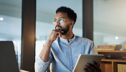 A young man rests his hand on his chin and looks at a laptop computer screen while holding a notebook in an office