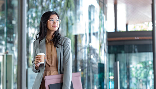 A young woman holds a coffee cup and briefcase outside a glass-walled building