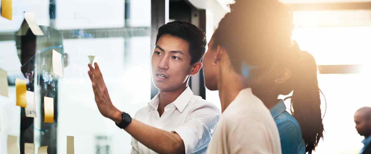 A young man gestures towards a clear dry erase board with sticky notes while speaking with female colleagues