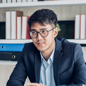 A man in a suit and glasses smiles in front of a bookcase.