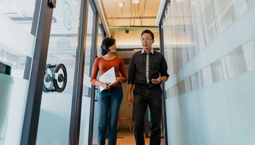 A man and woman talk together as they walk down a brightly lit office hallway.