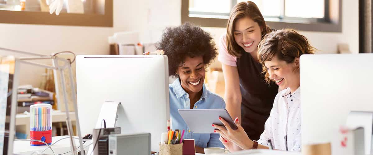 Three women smile and look at a tablet computer behind an office desk.