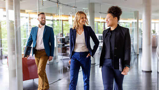 Three smiling coworkers in suits walks down a brightly lit office hallway.