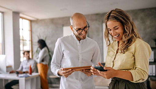 A smiling man and woman compare mobile devices in a brightly lit office.