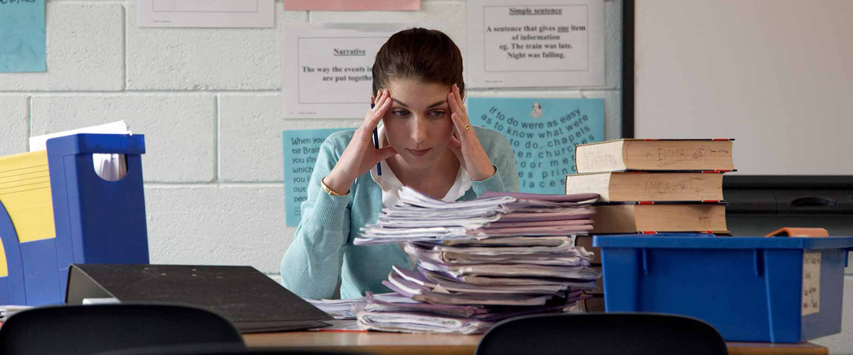 A female teacher sits behind a tall stack of papers and rests her hands on her temples.