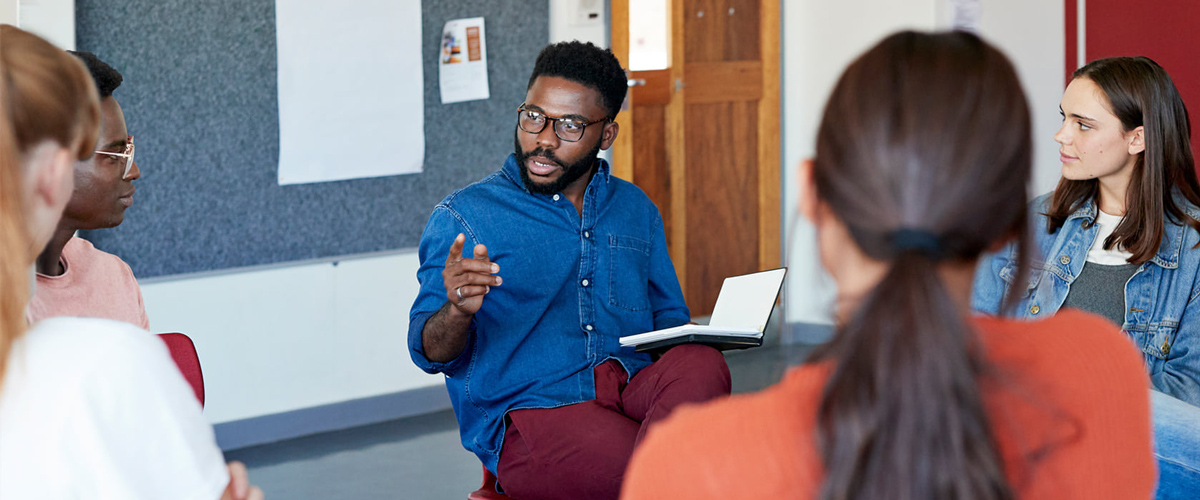 A teacher speaks and gestures with his hand while students listen as they sit in a circle in a classroom.