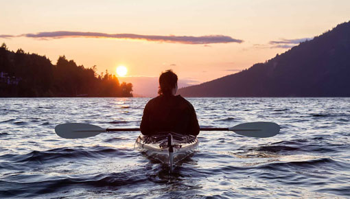 A kayaker sits on a placid lake with a sunset in the distance.