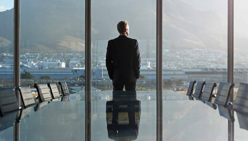 A man in a suit stands with his back to an empty boardroom as he looks out a large window.