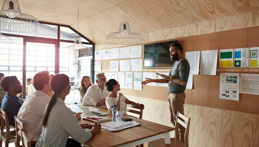 A group of coworkers listens to a male colleague speak in front of a wall with charts