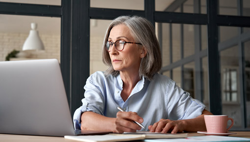 An woman with gray hair and glasses works at a laptop while writing in a notebook