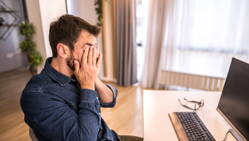 A young man rubs his eyes in front of his computer, seated at a desk