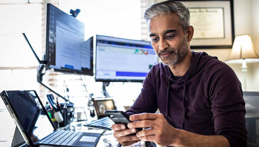 Man looks at smart phone while sitting at office desk with multiple computer monitors