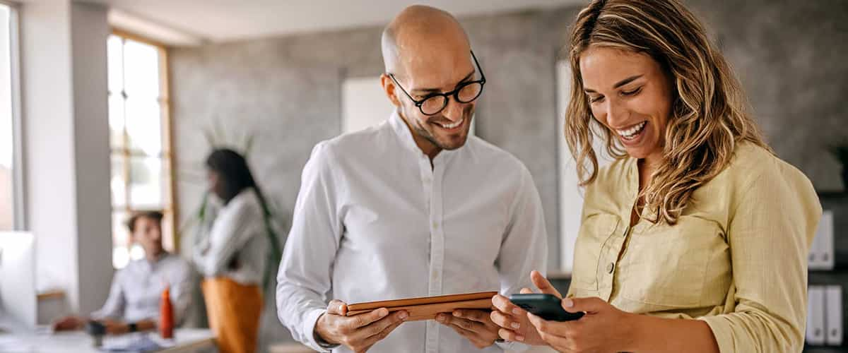 Two coworkers smile and look at mobile devices in a brightly lit office.