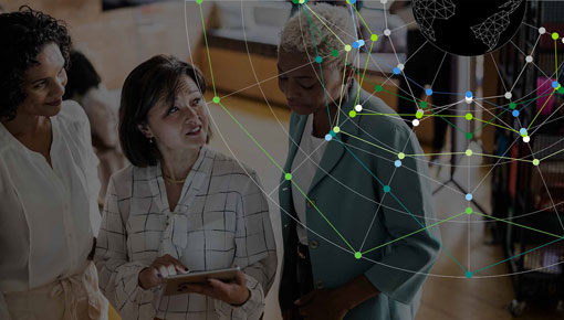 Three women in business attire look at a tablet computer in an office, with an overlay of a brightly lit network graphic.