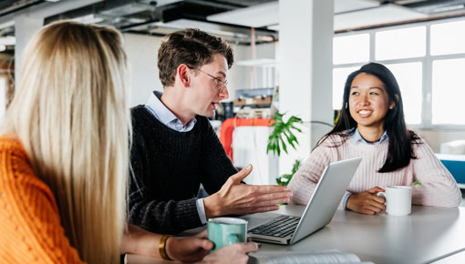 A mixed race group of coworkers talk together in an office in front of an open laptop computer