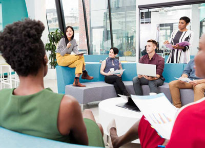 A group of coworkers gather in a circle on couches with laptops and papers.