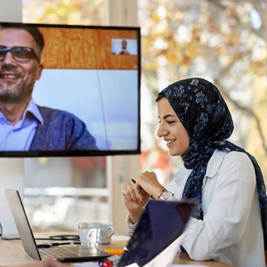 A smiling woman in a headscarf sits in front of a laptop computer with a colleague on video conference on a screen behind her.