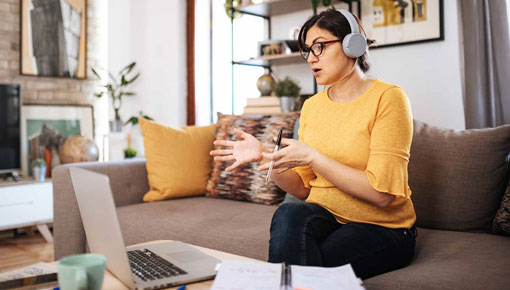 A young woman speaks to a video conference on her laptop while wearing headphones in her home office.