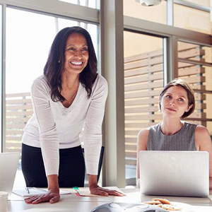 A smiling business woman leans over a table next to another woman sitting behind a laptop computer.