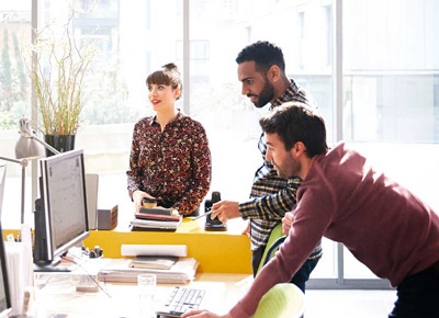 A group of coworkers gather around a desk with a computer monitor in a brightly lit office