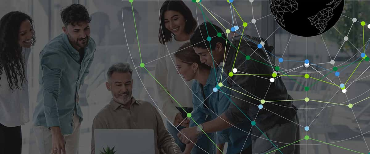 A group of coworkers smile and talk around a table with a laptop computer, covered by a black overlay with a globe and brightly lit network superimposed on top.