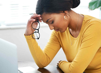 A young woman presses her hand to her forehead, while holding her glasses, in front of a laptop 