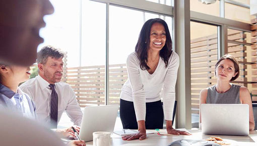 Smiling woman in business attire leans over a board room table with a group of coworkers