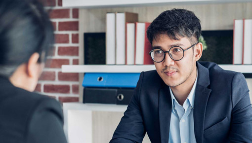 An Asian man in a suit and glasses smiles at a colleague in an office