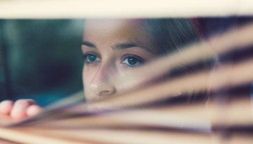 Woman looks warily through blinds out a window