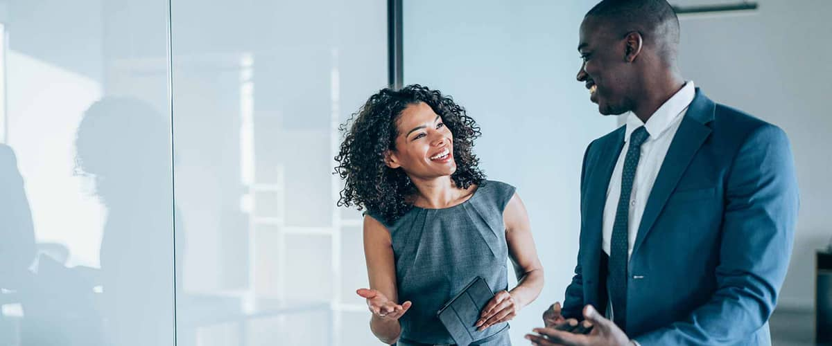 Two smiling business people talk while walking in a hallway.