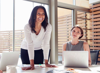 A smiling woman leans over a table next to a young woman with a laptop computer 