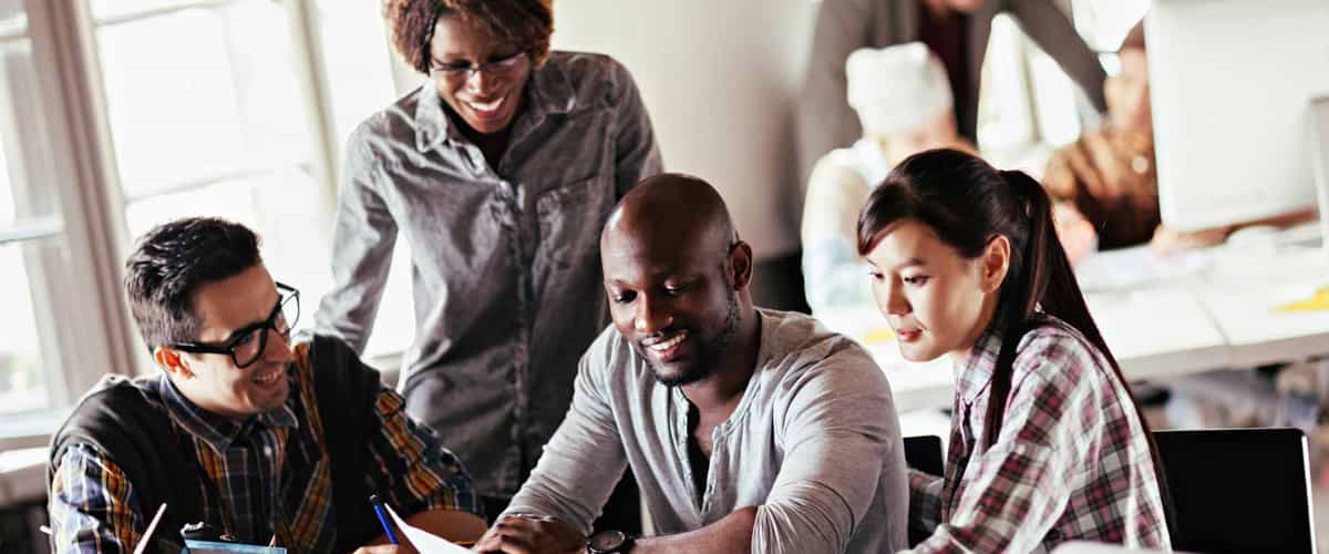 A group of students sit at a table, smiling and looking at a piece of paper, while a teacher stands behind them.