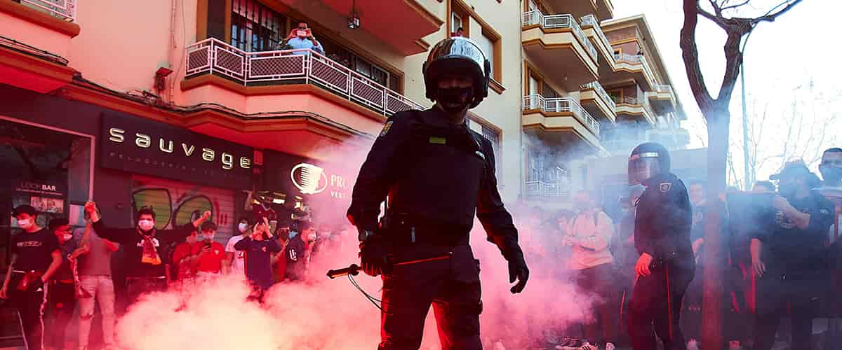 Police man in armor standing in a red swirl of gas and surrounded by a crowd. 