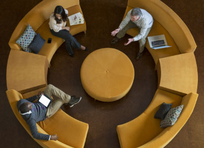 A woman and two men sitting on a cirular yellow couch having a conversation.