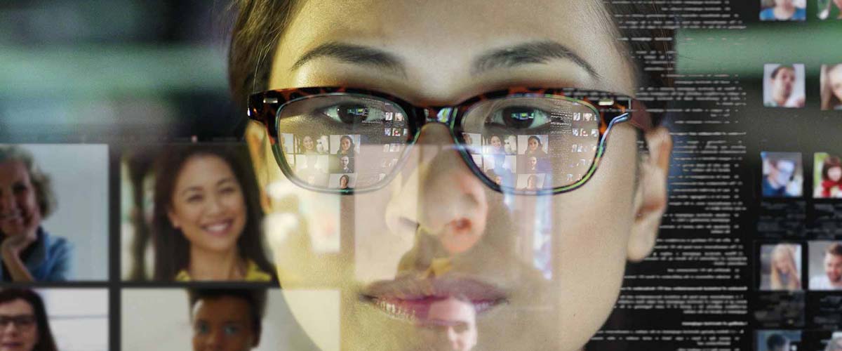 A woman with glasses reflected in a computer screen with many Zoom meetings/video conferences open.