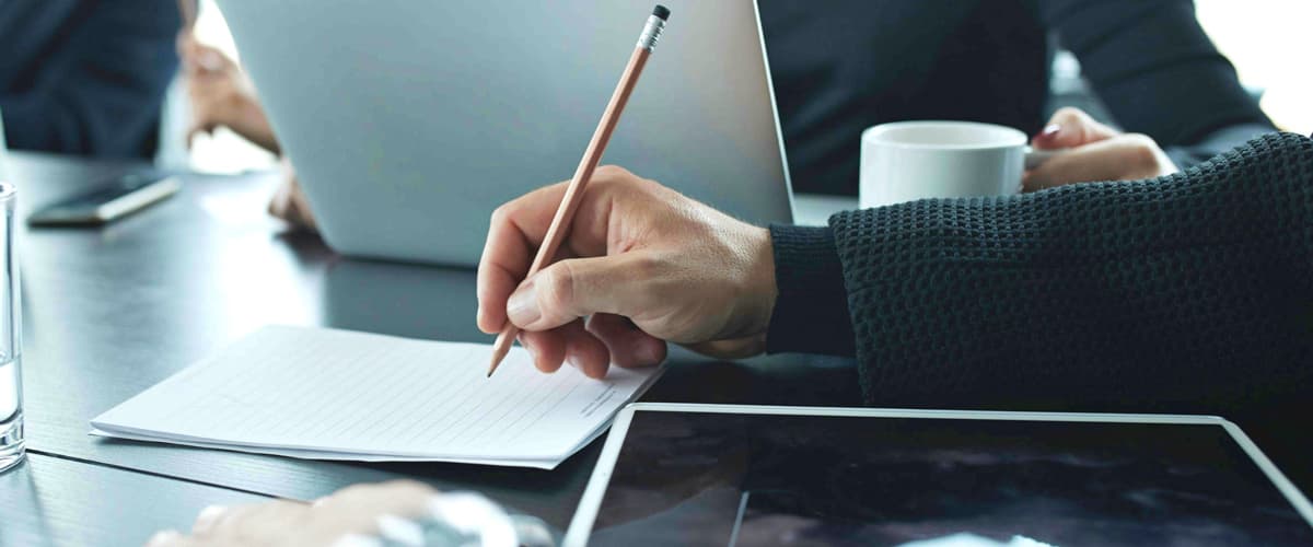 Image of a person holding a pencil above a stack of papers in a office setting with others