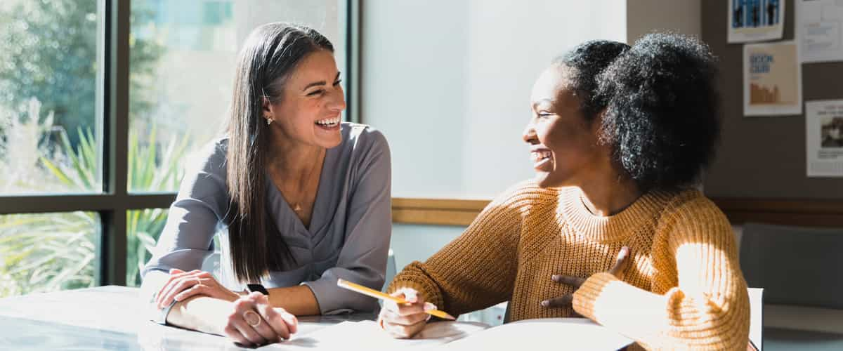 A female teacher tutors a teenage girl after school. They are smiling as they look at each other